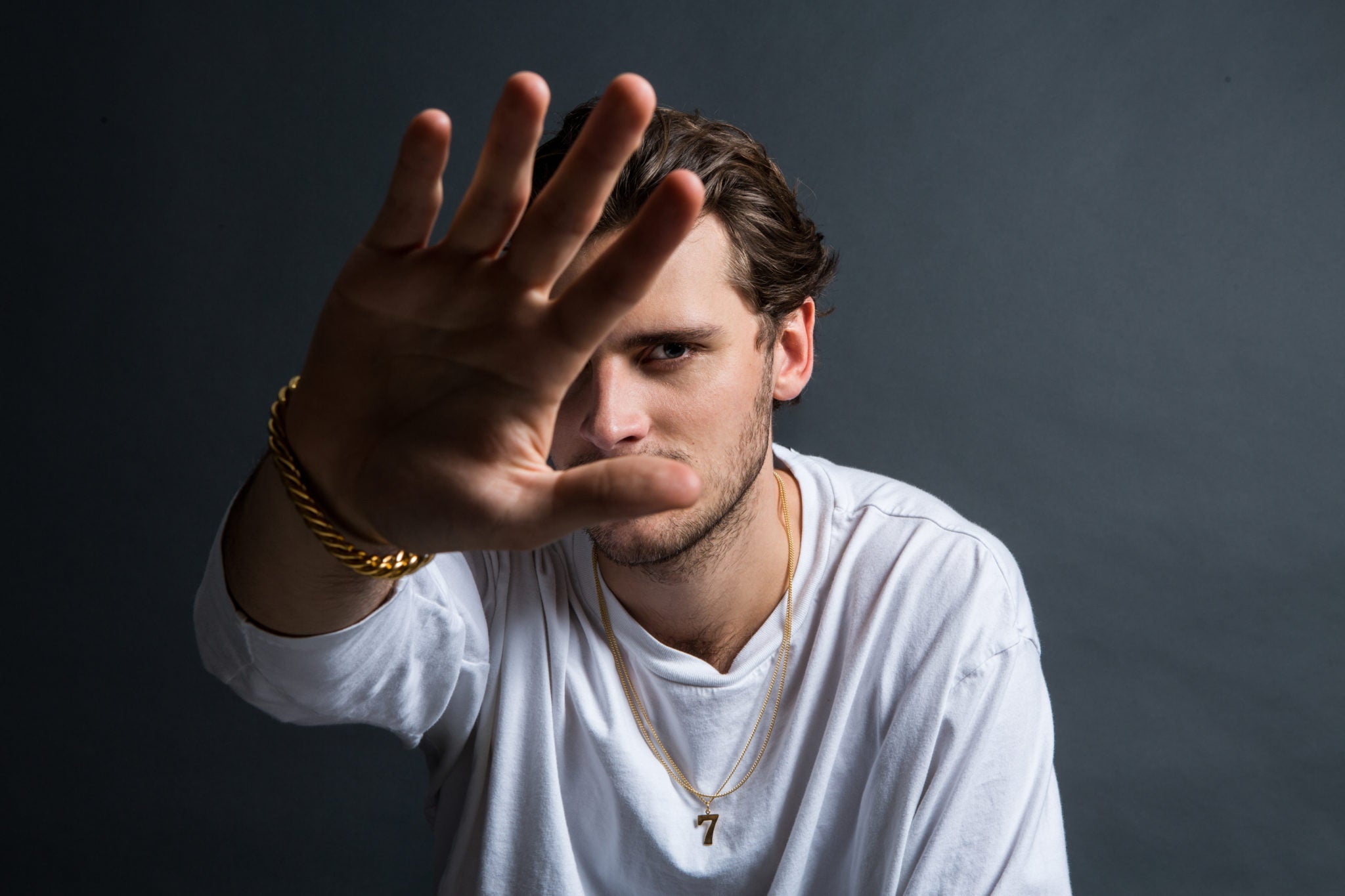 Man in a white shirt with gold jewellery making a stop gesture with his hand against a dark background - Tilly Sveaas